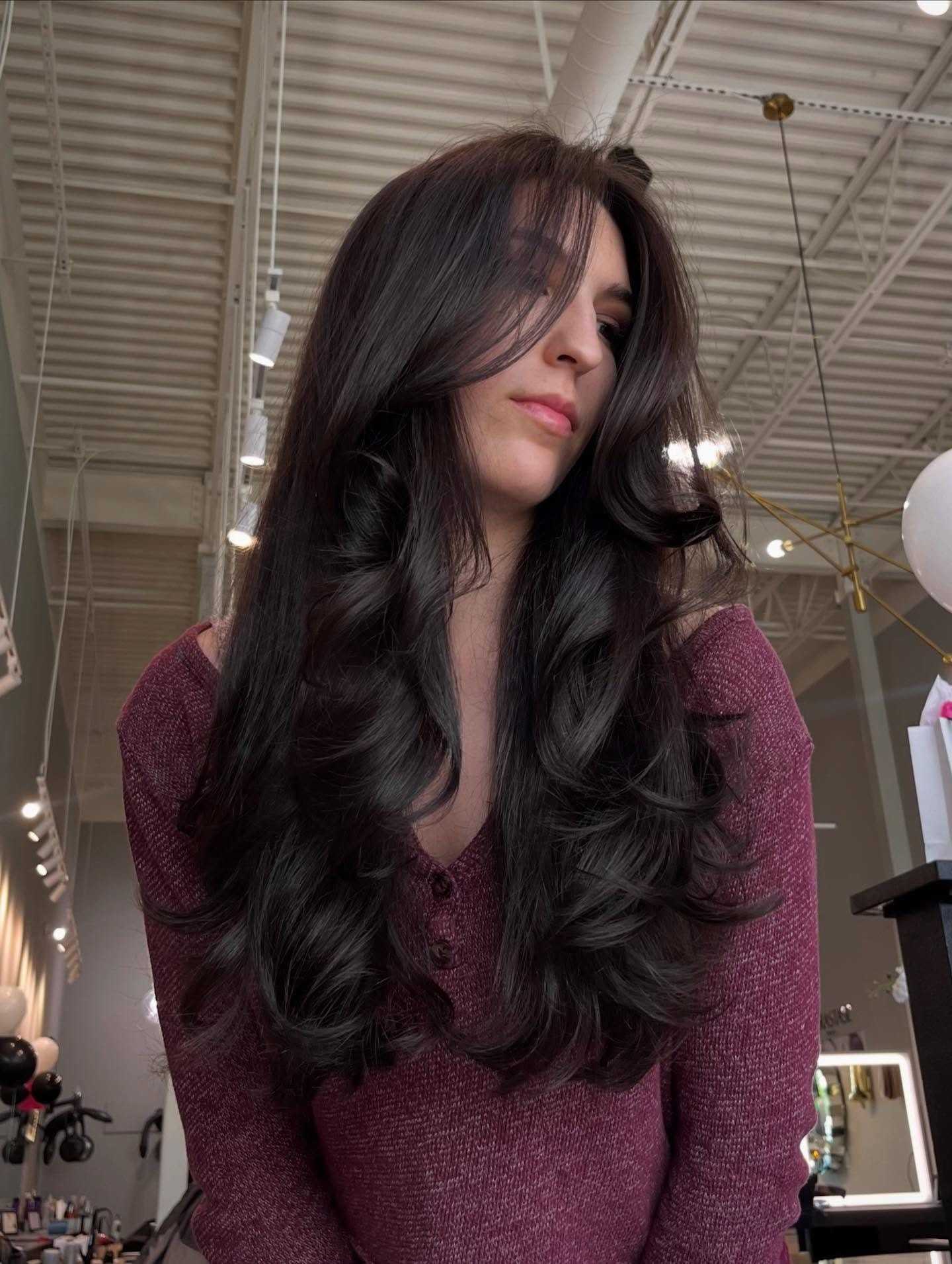 Young woman with long, wavy, dark hair in a salon setting, wearing a maroon top.