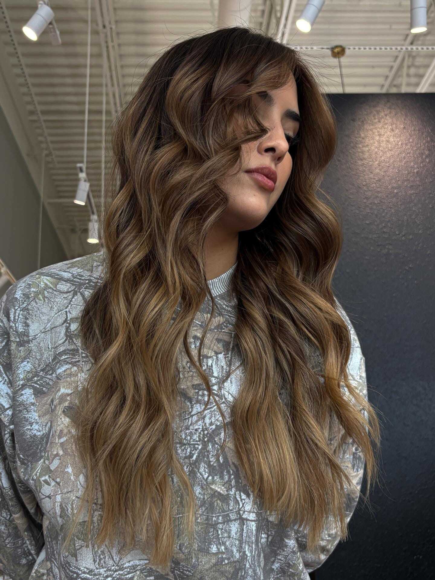 Woman with long, wavy brown hair wearing a patterned sweater in a well-lit room.