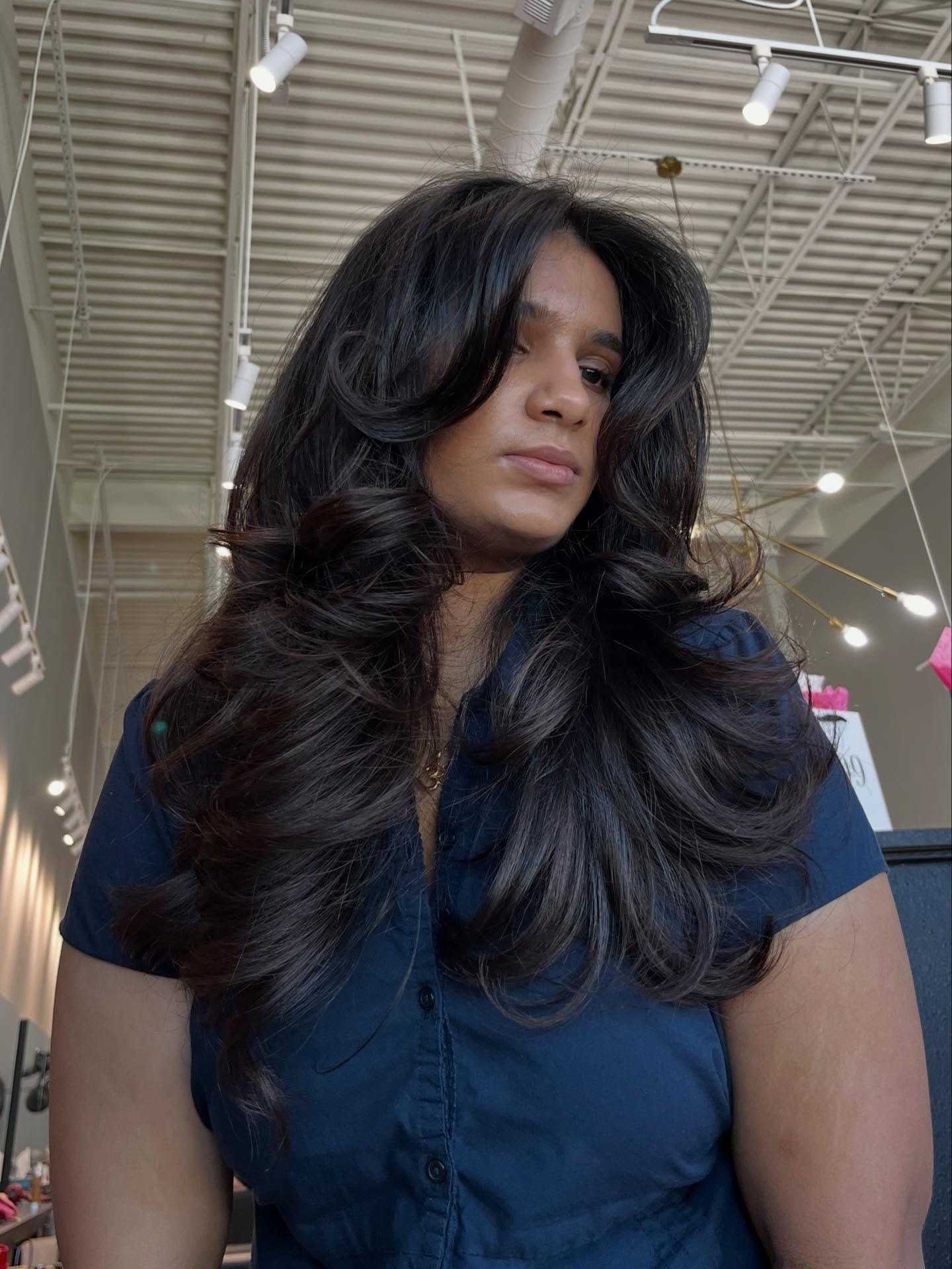 Woman with long, dark, wavy hair in a salon, wearing a navy shirt.