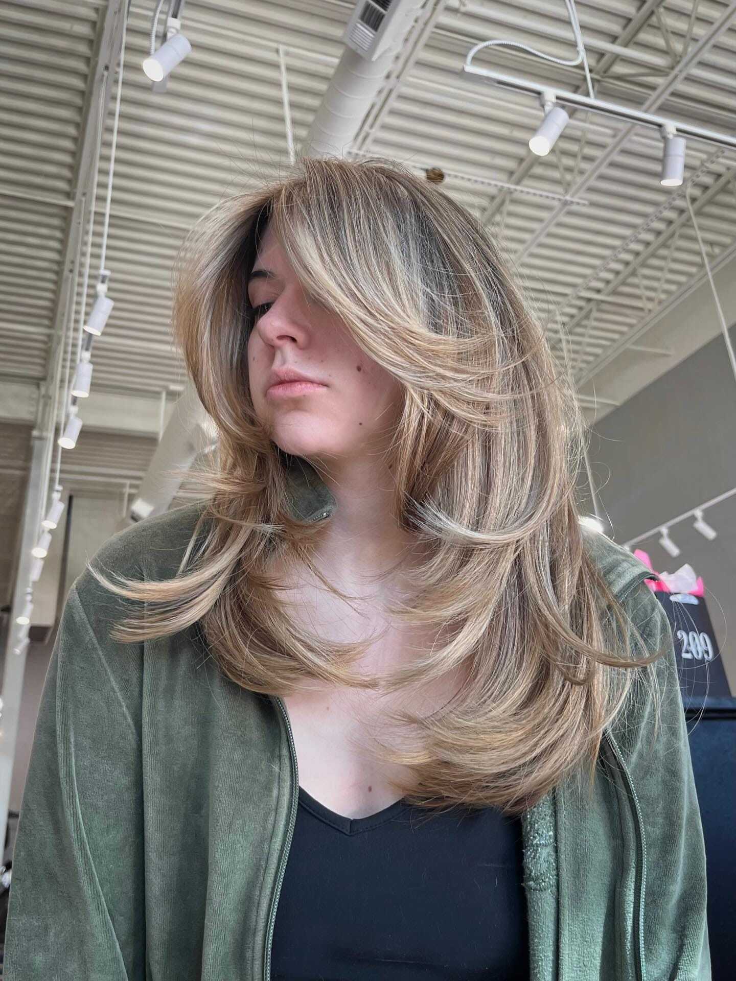 Woman with layered blonde hair in a salon, wearing a green jacket.
