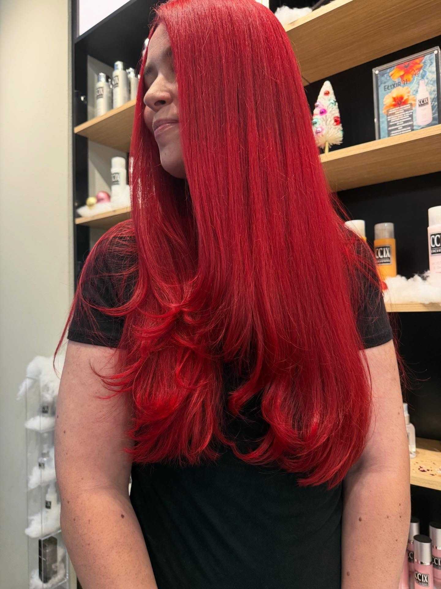 Woman with vibrant red hair smiling in a salon, surrounded by hair products.