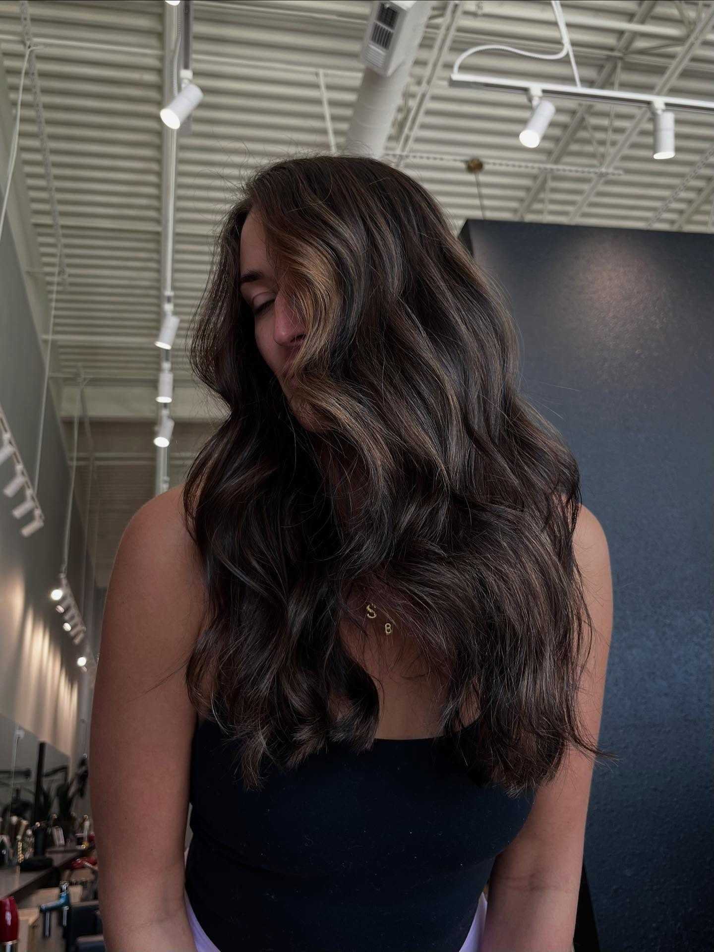 Woman with long, wavy brown hair, wearing a black top, in a modern interior space.