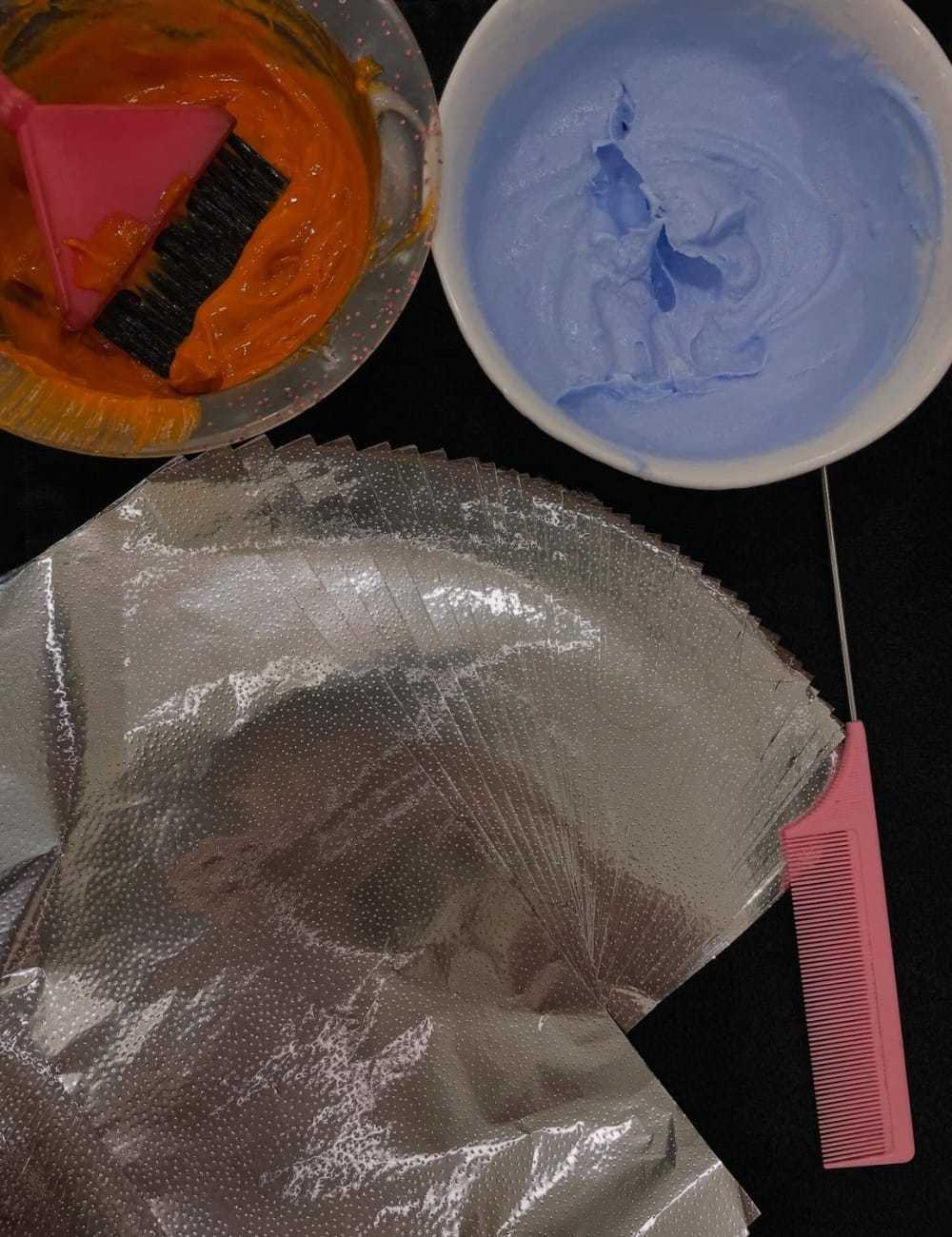 Hair coloring tools with orange and blue dye, foils, brush, and comb on a black surface.