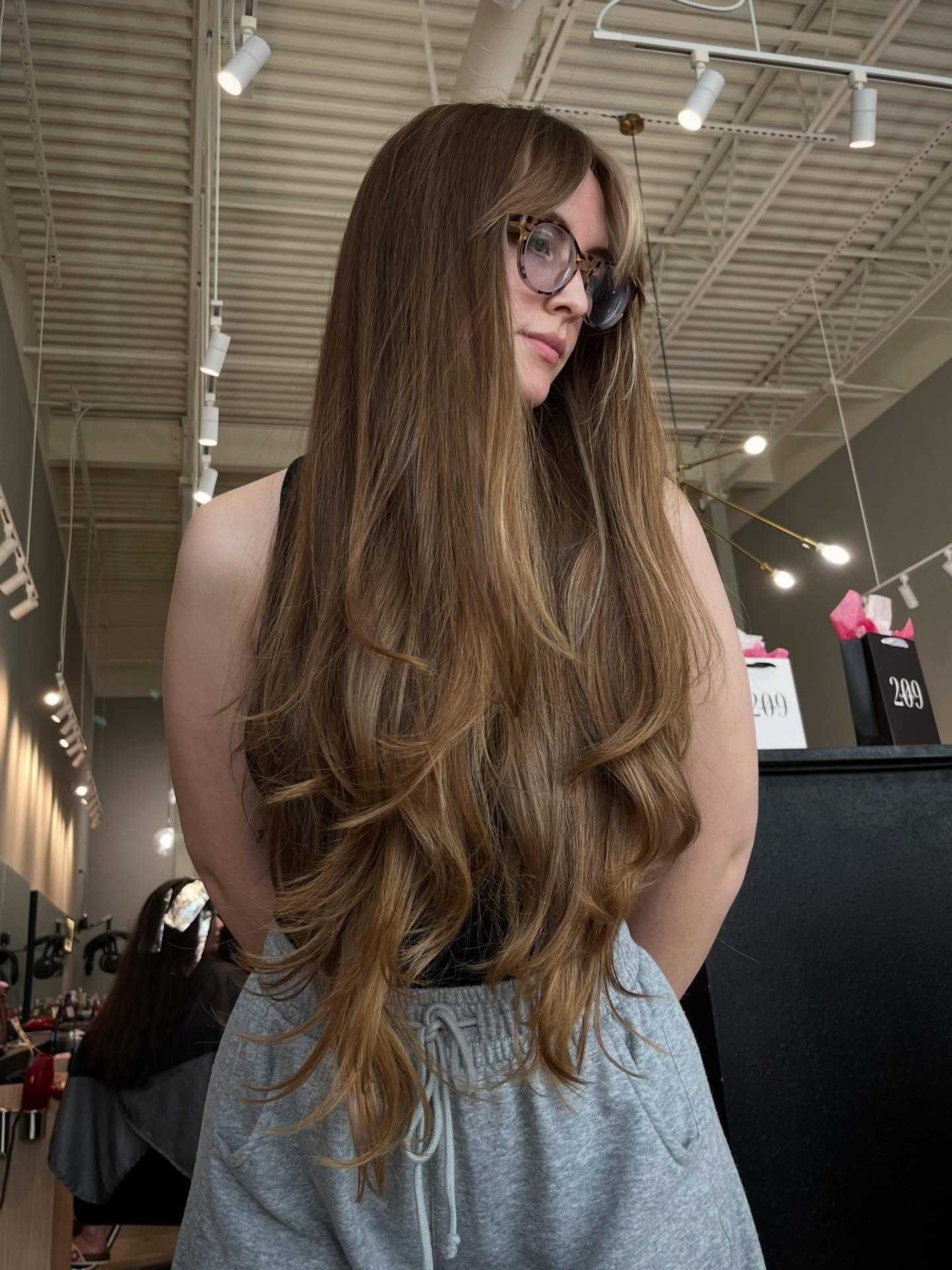Long-haired woman in salon with stylish glasses, gray pants, and black top.
