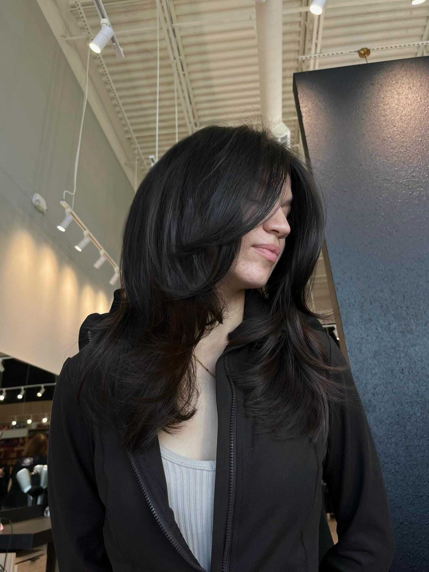 Woman with long, dark layered hair in a salon, wearing a black jacket.